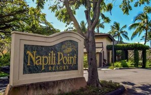 Schild für das Napili Point Resort mit freiem Blick auf die umliegende Bepflanzung und einem Gebäude im Hintergrund.