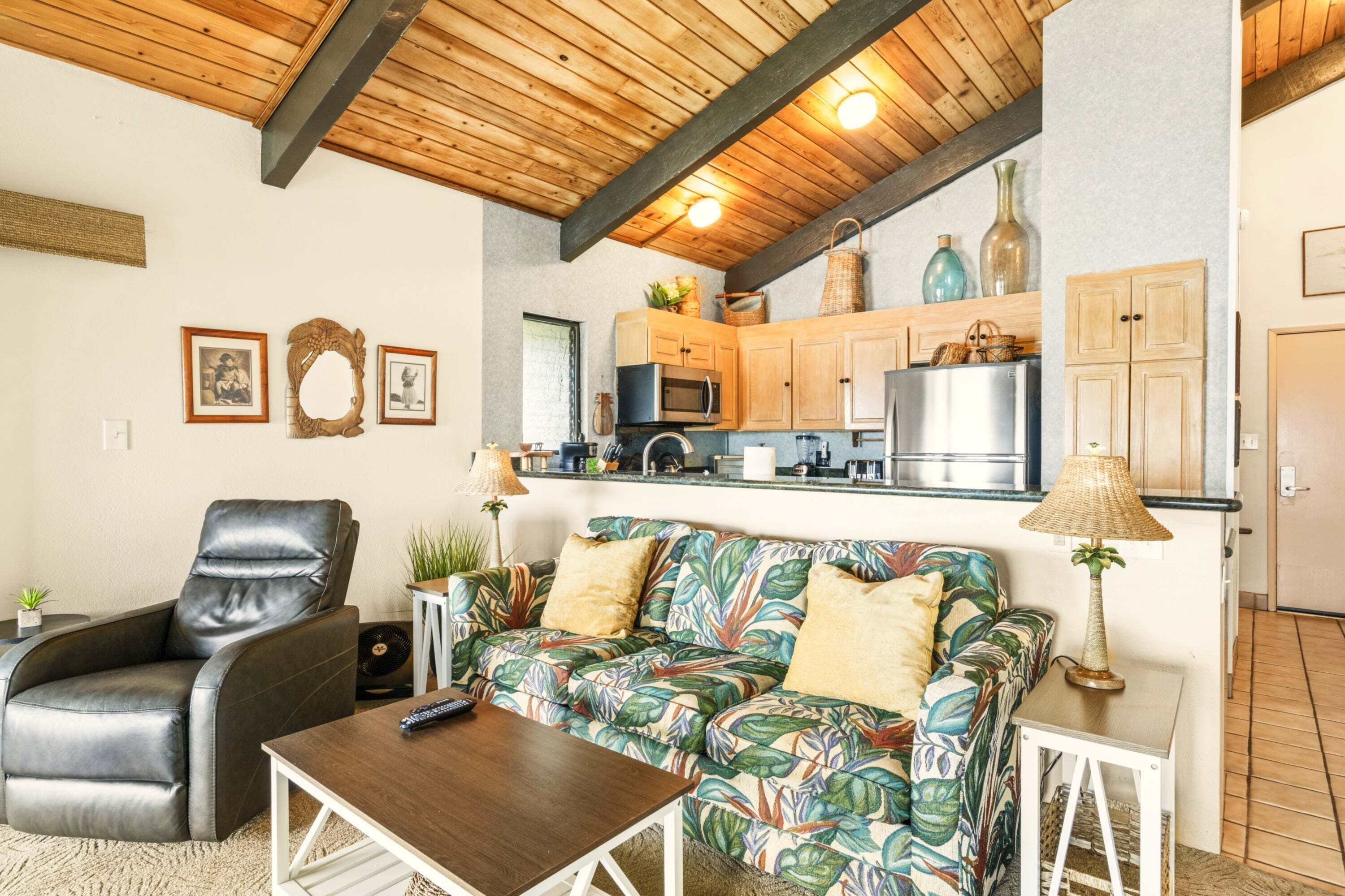 Un salon chaleureux avec un fauteuil en cuir, un canapé à motifs tropicaux, une table basse et un coin cuisine doté de meubles en bois et d’un réfrigérateur.