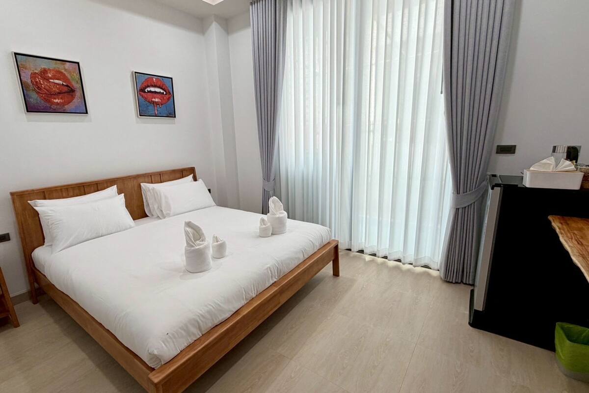 A hotel room with a wooden bed, white bedding, two framed artworks on the wall, a desk with a chair, and a window with sheer curtains.