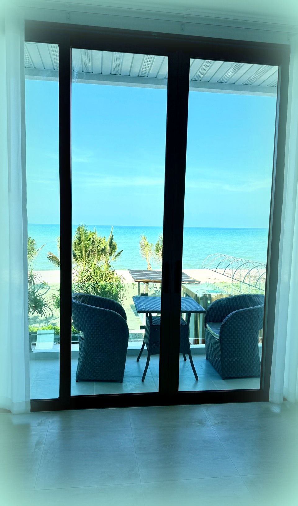A room with a large sliding glass door leading to a balcony with a view of the ocean and palm trees.