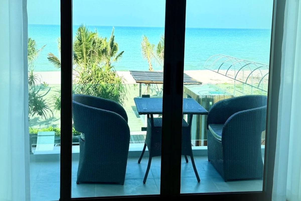 Una habitación con un amplio ventanal corredizo que da a un balcón con vistas al océano y palmeras.