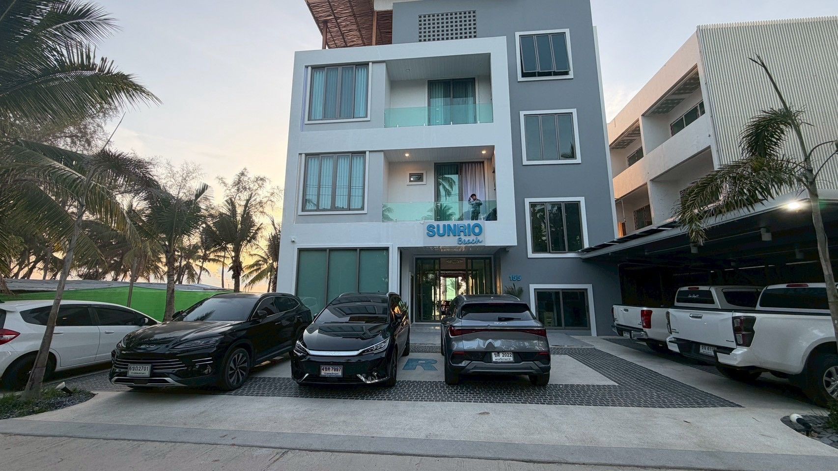 A multi-story building with a commercial establishment on the ground floor named 'SUNRIO Beach.' There are several cars parked in front, and the building has a modern architectural style with large windows and balconies.