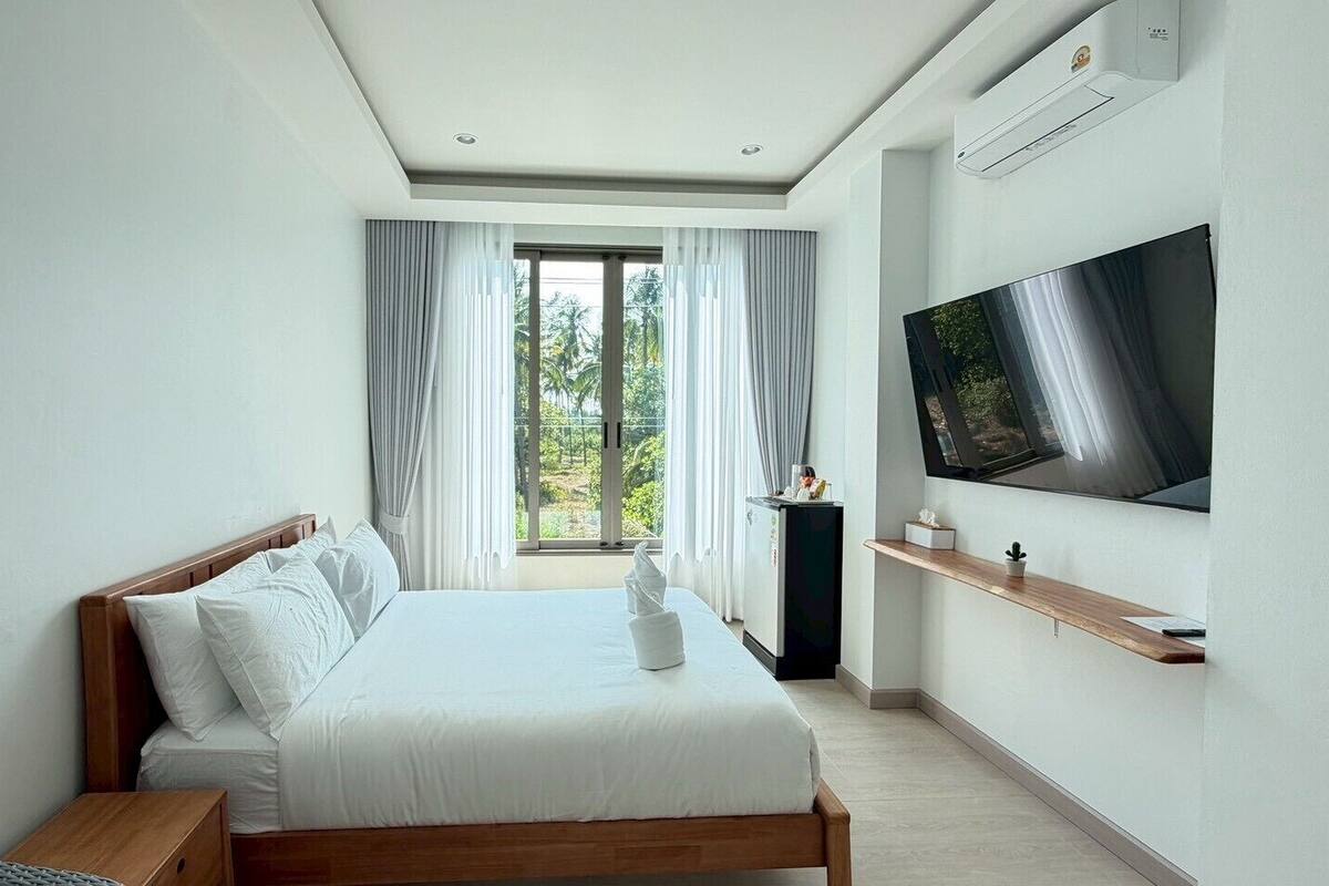 A modern hotel room with a large bed, a flat-screen TV, a small table, and a view of greenery through the window.