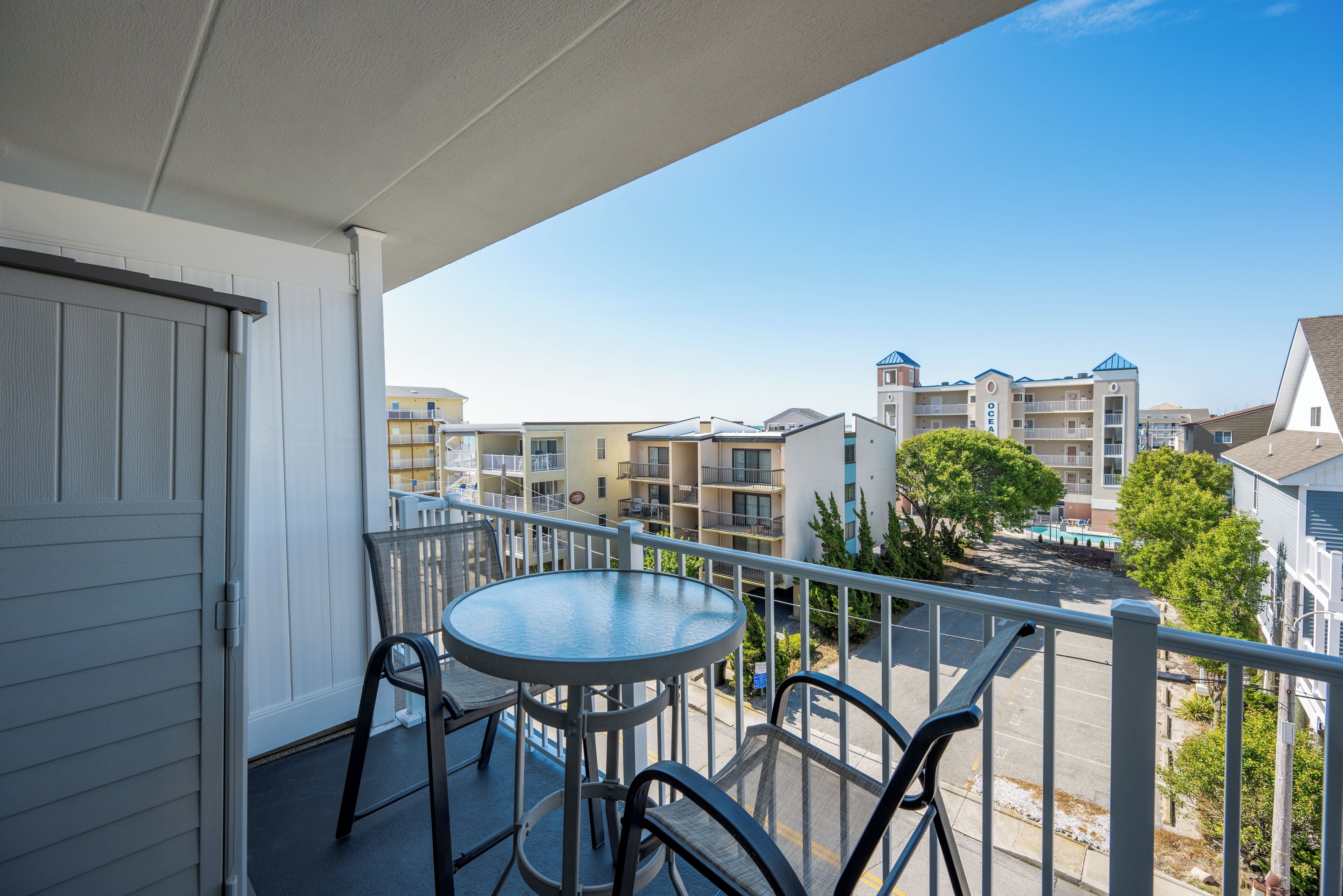 A balcony with a table and chairs, overlooking a residential area with multiple buildings and a clear sky.
