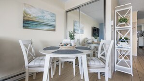 A dining area with a round table, white chairs, a white shelving unit with plants, and a painting of a seascape. - Just listed for rent! Oceanview condo in an oceanfront building with a pool, elevator and assigned parking (Ocean City)