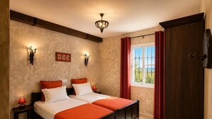 A bedroom with two beds, a window with curtains, wall sconces, and a wooden ceiling beam.