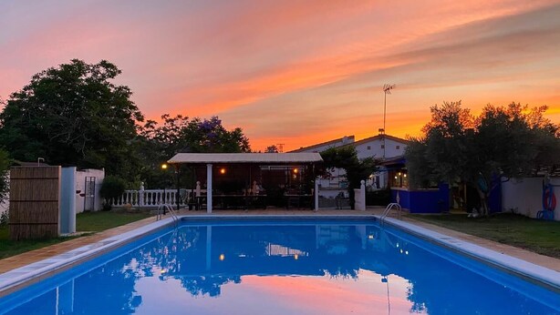 Ein Pool mit einer Sonnenterrasse, umgeben von Bäumen und einem Gebäude mit überdachter Sitzgruppe.