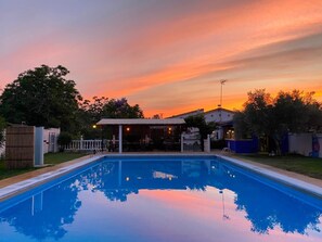 A swimming pool with a patio area, surrounded by trees and a building with a covered seating area.