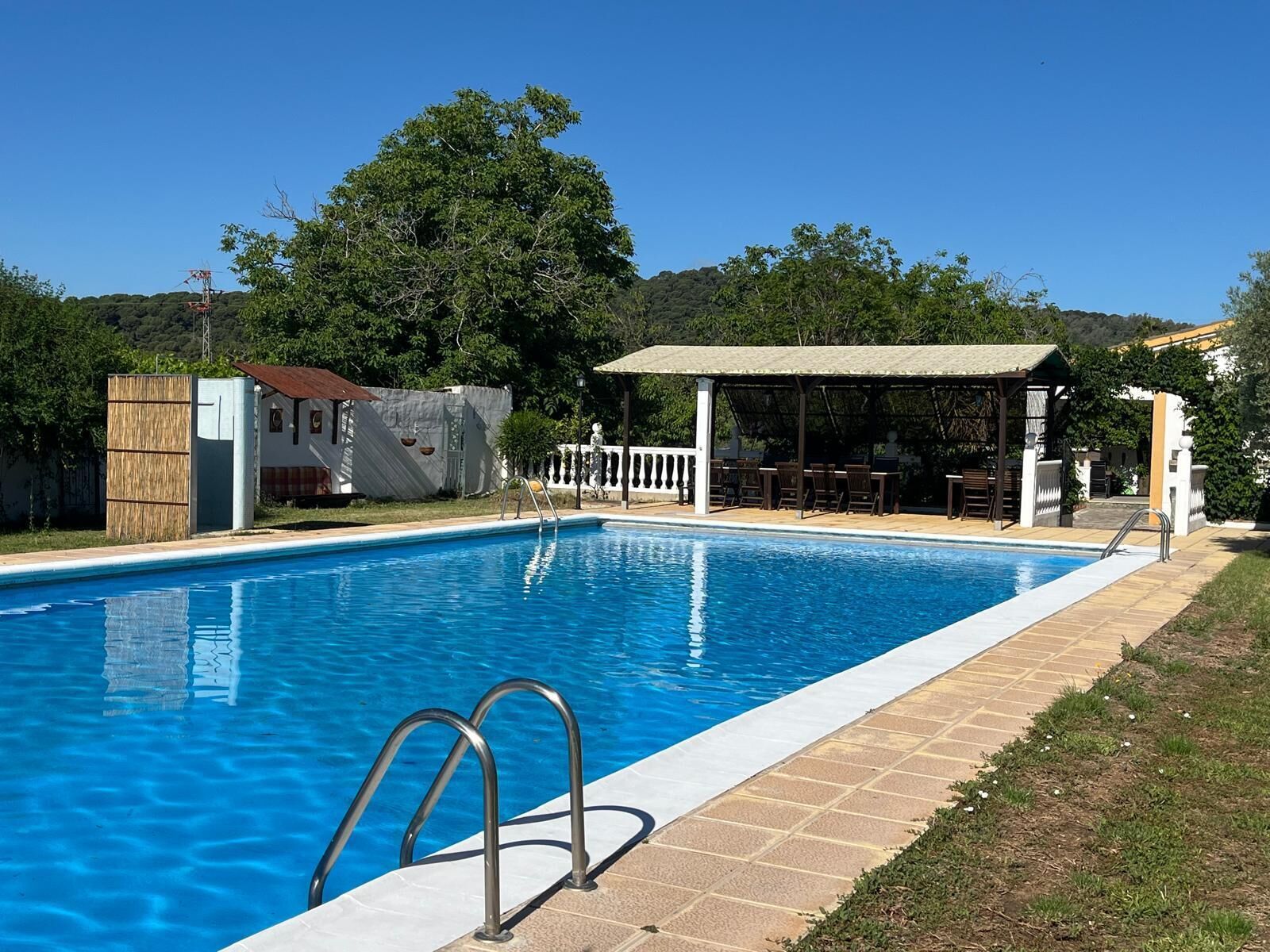 Una piscina amb una escala metàl·lica, zona de seients coberta i jardí tancat per una tanca.