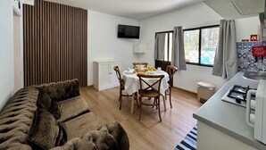 Een kamer met een bank, een eettafel met stoelen, een aan de muur bevestigde televisie, een raam met gordijnen en een keukenhoek met een spoelbak en fornuis.