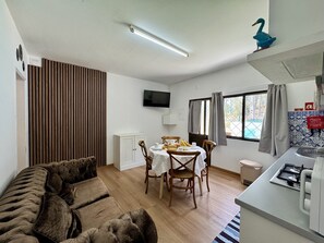 Une pièce comprenant un canapé, une table à manger avec des chaises, un téléviseur fixé au mur, une fenêtre avec des rideaux et un coin cuisine avec un évier et une cuisinière.