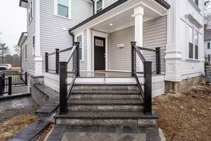 Ein zweistöckiges Haus mit Veranda, schwarzem Metallgeländer und Steinstufen, die zur Tür führen.