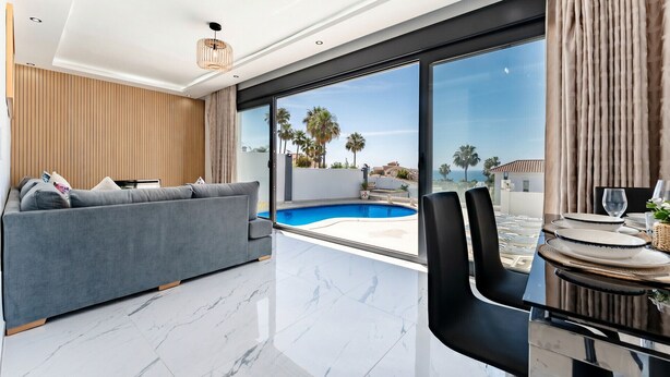 Een moderne woonkamer met een grijze bank, een eethoek met een tafel en stoelen, een groot raam met uitzicht op een zwembad en palmbomen, en een plafond met inbouwspots.