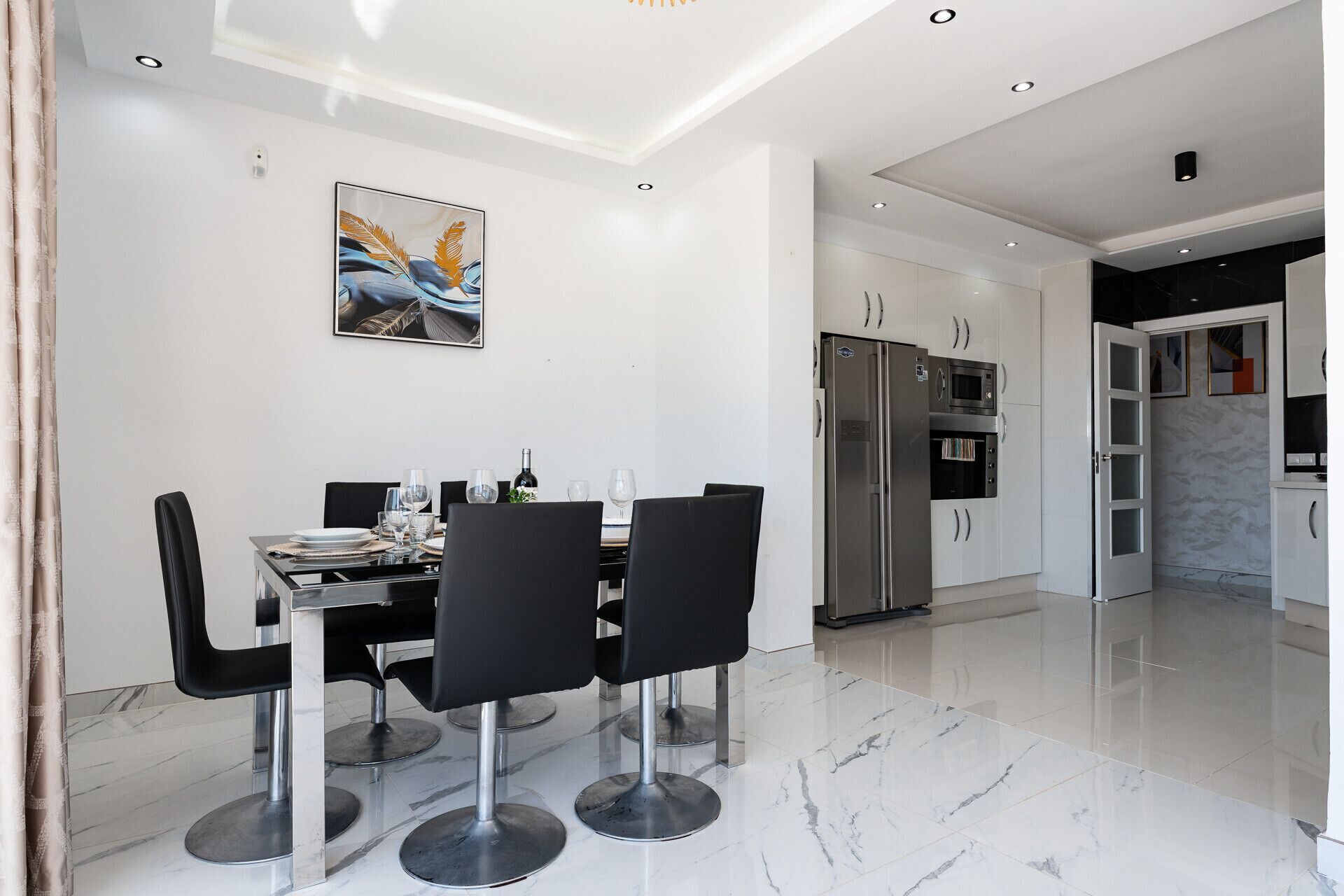 A modern dining area with a marble floor, a dining table with chairs, a kitchen with appliances, and a painting on the wall.