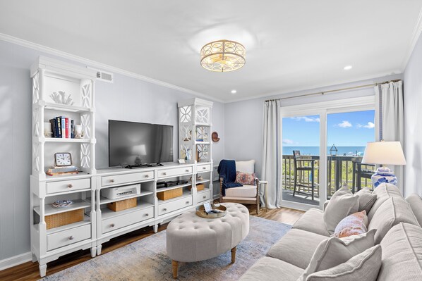 A living room with a TV, white entertainment unit, ottoman, sofa, and a view of the sea through large windows. - Chateau By The Sea-Oceanfront-Sleeps4-2b/2b-King Beds-Laundry-Kitchen (North Myrtle Beach)