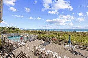 A wooden deck with pool, chairs, and tables overlooking a beach. - Chateau By The Sea-Oceanfront-Sleeps4-2b/2b-King Beds-Laundry-Kitchen (North Myrtle Beach)