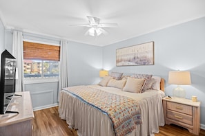 A bedroom with a bed, bedside lamps, a ceiling fan, a window with blinds, and a wall-mounted artwork. - Chateau By The Sea-Oceanfront-Sleeps4-2b/2b-King Beds-Laundry-Kitchen (North Myrtle Beach)