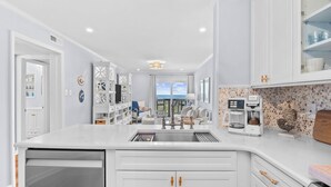 A modern kitchen with white cabinetry, a built-in dishwasher, a coffee maker, and a view to the outside. - Chateau By The Sea-Oceanfront-Sleeps4-2b/2b-King Beds-Laundry-Kitchen (North Myrtle Beach)