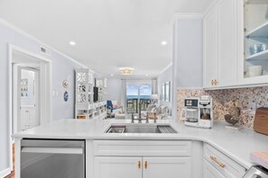 A modern kitchen with white cabinetry, a built-in dishwasher, a coffee maker, and a view to the outside. - Chateau By The Sea-Oceanfront-Sleeps4-2b/2b-King Beds-Laundry-Kitchen (North Myrtle Beach)