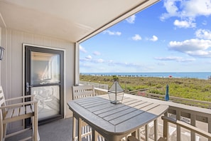 A balcony with a table and chairs, overlooking a beach with a clear sky. - Chateau By The Sea-Oceanfront-Sleeps4-2b/2b-King Beds-Laundry-Kitchen (North Myrtle Beach)