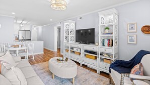 A modern living room with a TV, white furniture, and a dining area. - Chateau By The Sea-Oceanfront-Sleeps4-2b/2b-King Beds-Laundry-Kitchen (North Myrtle Beach)