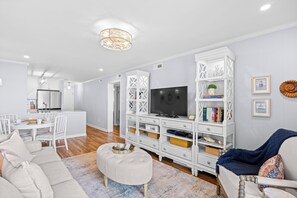 A modern living room with a TV, white furniture, and a dining area. - Chateau By The Sea-Oceanfront-Sleeps4-2b/2b-King Beds-Laundry-Kitchen (North Myrtle Beach)