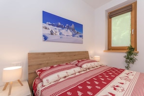 Een slaapkamer met een houten hoofdeinde, een bed met rode en witte beddengoed, een wanddecoratie van een besneeuwde berg, een raam met houten jaloezieën, en een bedlampje.