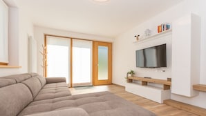 Ein modernes Wohnzimmer mit einem Ecksofa, einem Flachbildfernseher und einer Schiebetür zum Balkon.