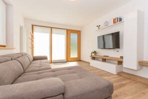 Ein modernes Wohnzimmer mit einem Ecksofa, einem Flachbildfernseher und einer Schiebetür zum Balkon.