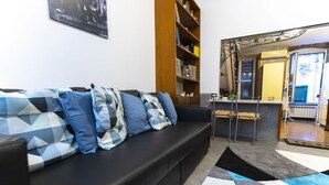 Una sala de estar con sofá negro, cojines estampados, una estantería de madera, una zona de cocina y un espejo grande.
