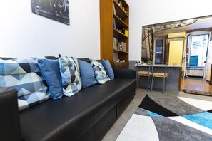 Ein Wohnzimmer mit schwarzem Sofa, gemusterten Kissen, einer Holzbibliothek und einer Küchenzeile, sowie einem großen Spiegel.