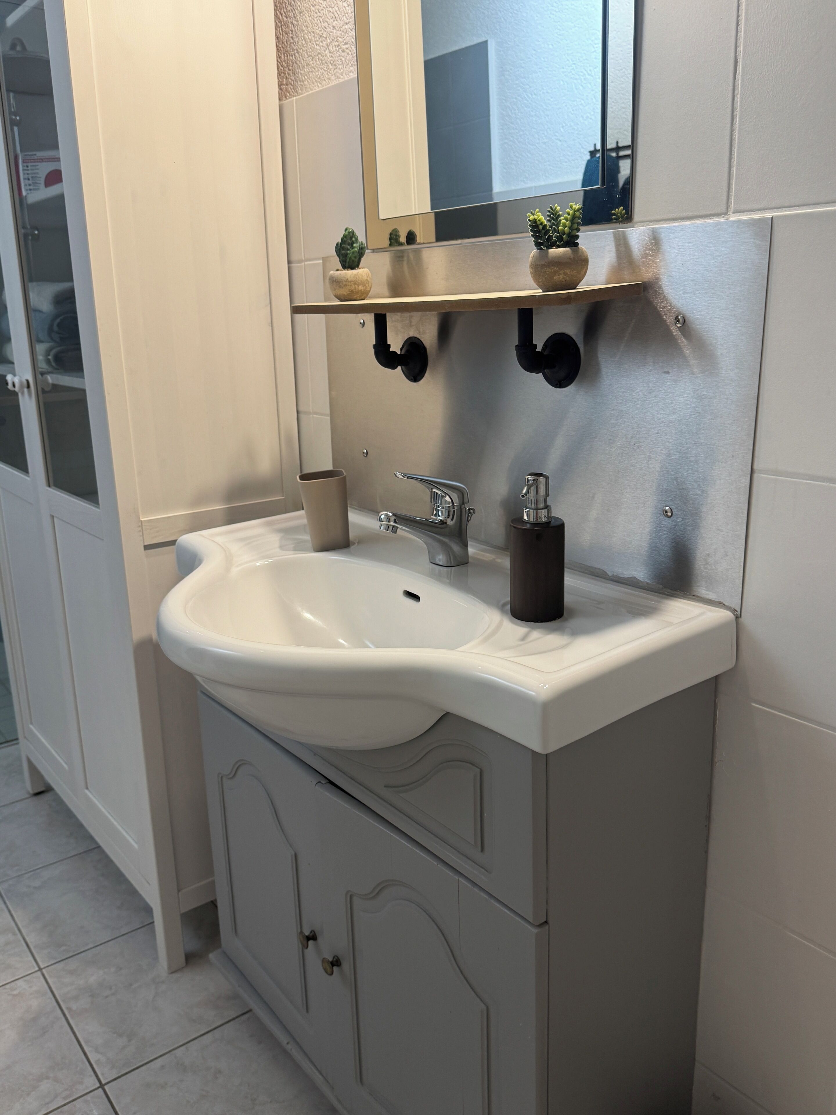 Un baño con un lavamanos blanco, un espejo, un estante con plantas pequeñas y un armario.