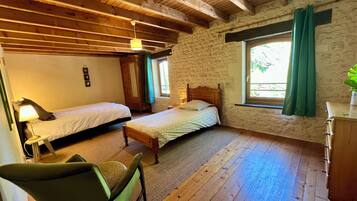 A room with two beds, a wooden ceiling, a stone wall, a window with curtains, and a chair.