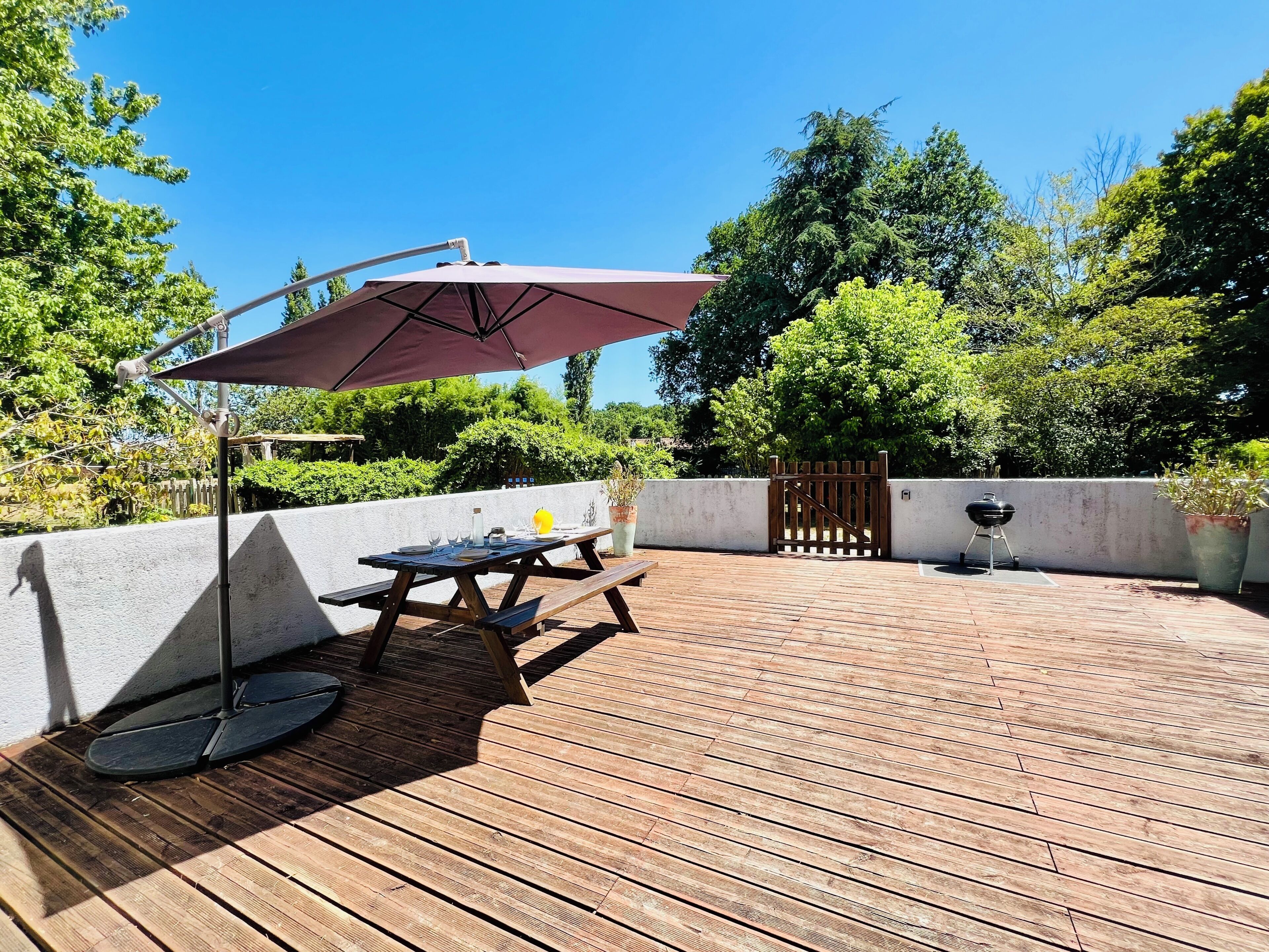 Une terrasse en bois avec une table de pique-nique et un parasol, entourée de verdure et agrémentée d’un barbecue.