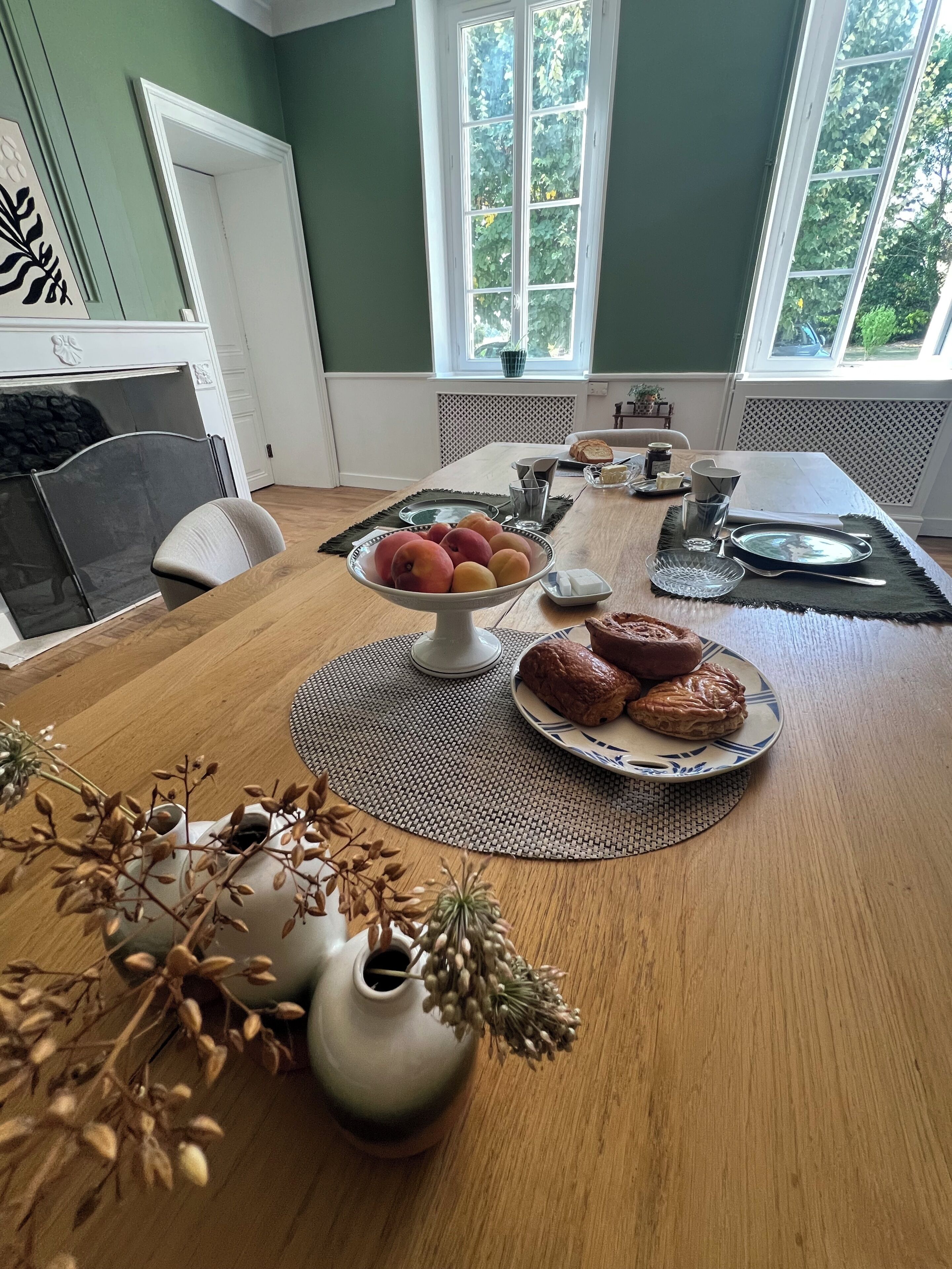 A dining table with a fruit bowl, a plate of pastries, and a vase with dried flowers.