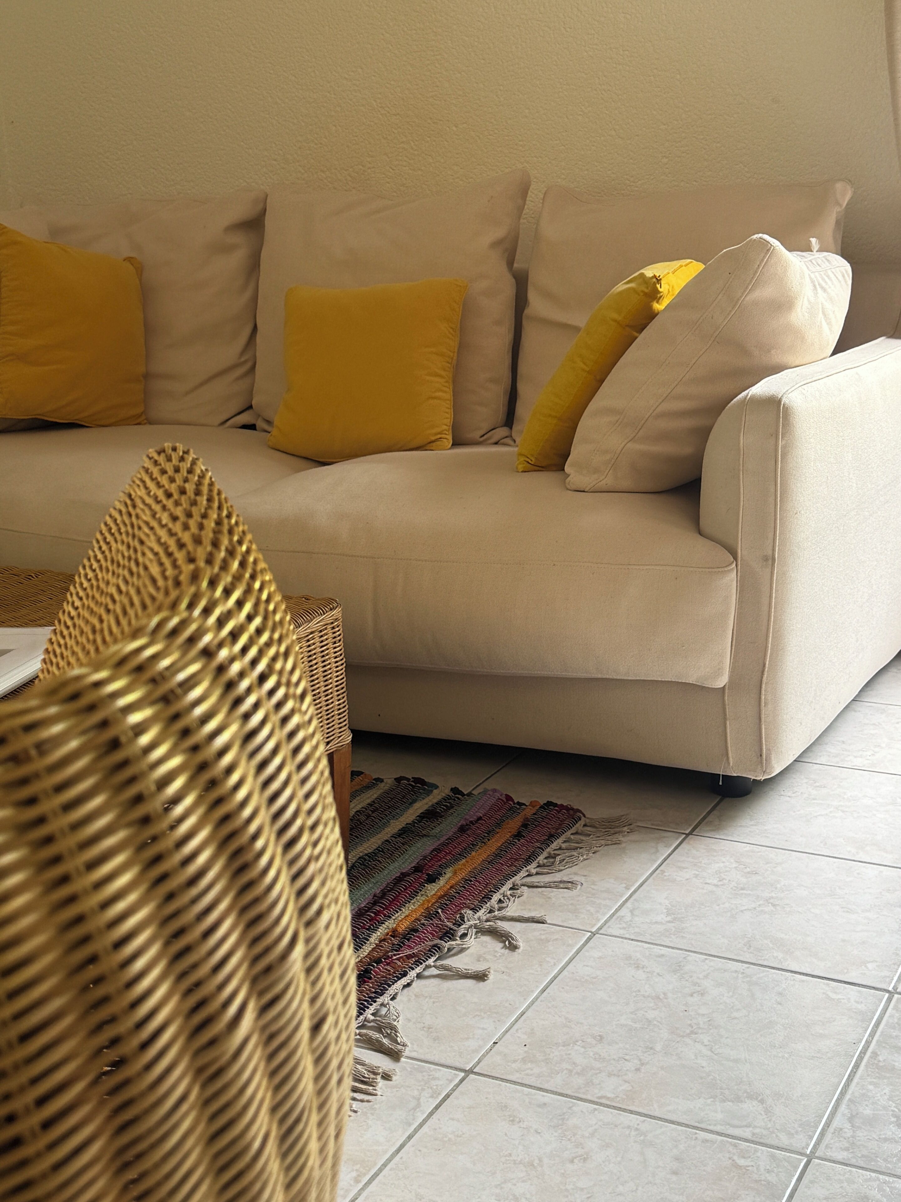 Un sofá color crema con cojines amarillos, una silla de fibra natural y una alfombra a rayas sobre un piso de baldosas.