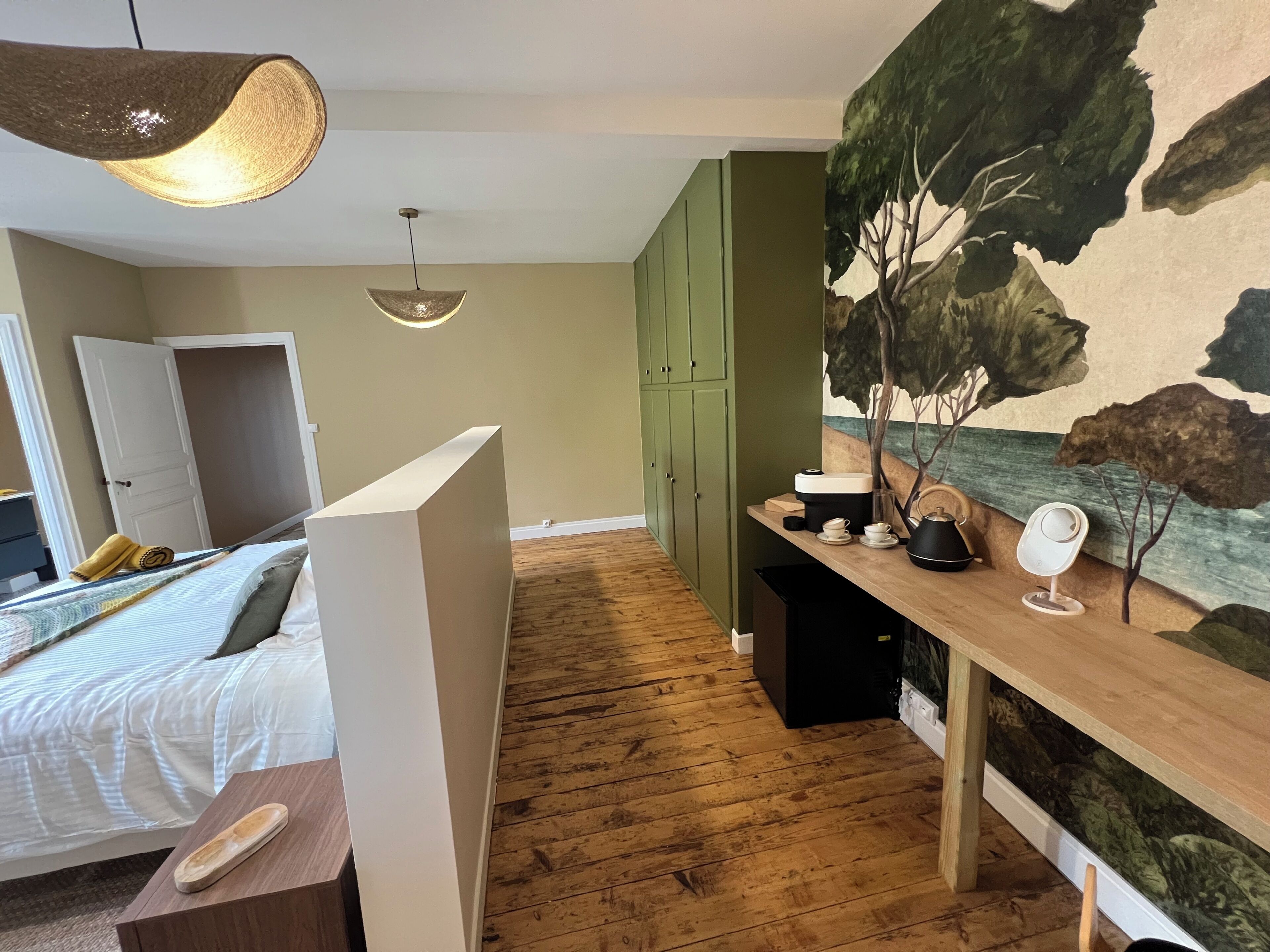 Quarto com uma cama, piso de madeira, guarda-roupa verde, escrivaninha com espelho e um quadro de uma árvore.