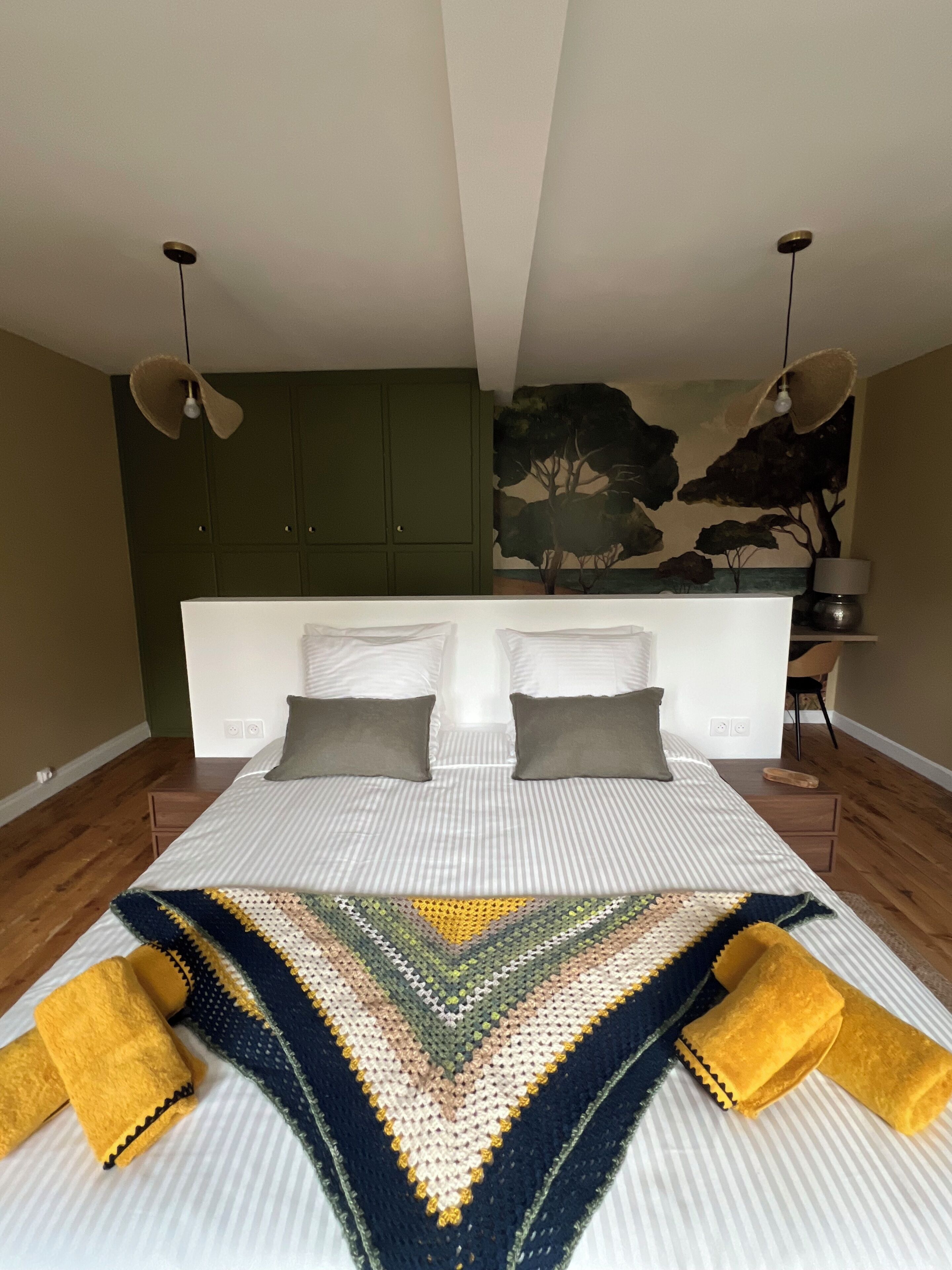 Une chambre à coucher avec un grand lit, deux oreillers, une tête de lit fixée au mur, un ventilateur de plafond et un tableau représentant des arbres.
