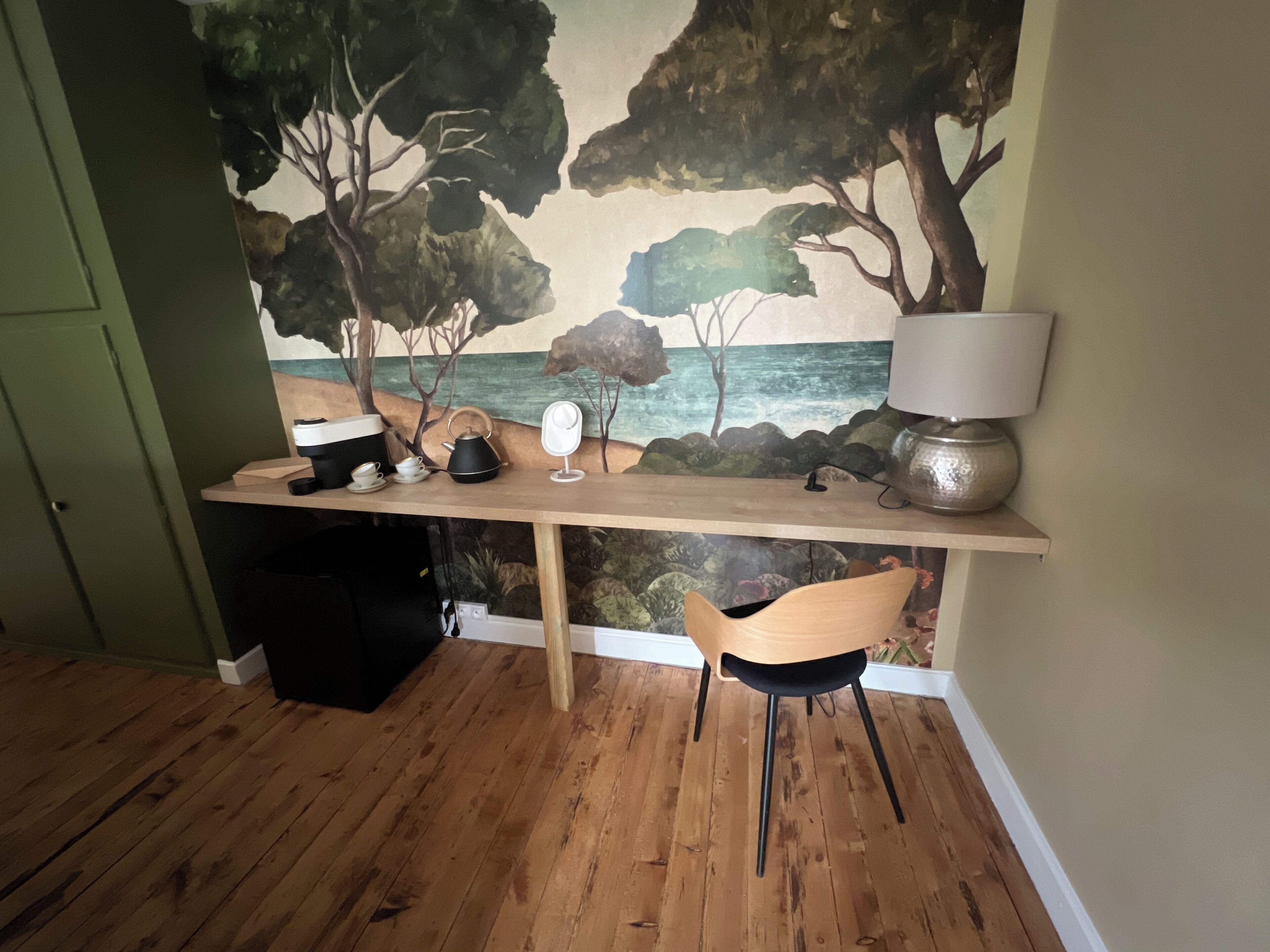 A room with a wooden desk, a chair, a lamp, and a mural of a coastal scene.