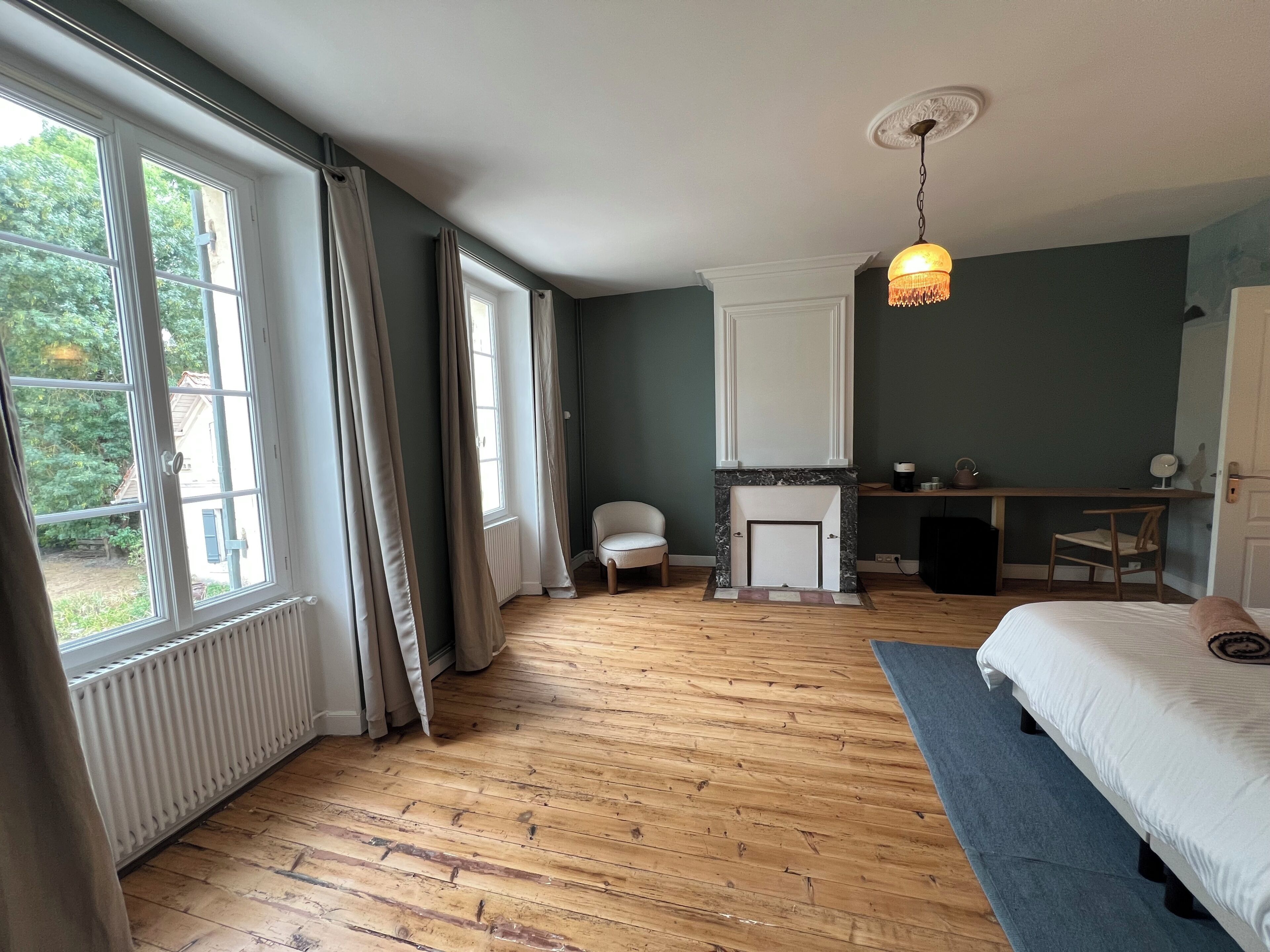 A bedroom with a wooden floor, a bed, a desk, a chair, a fireplace, and a window with curtains.