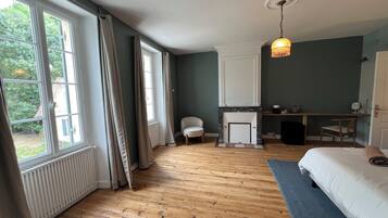 A bedroom with a wooden floor, a bed, a desk, a chair, a fireplace, and a window with curtains.
