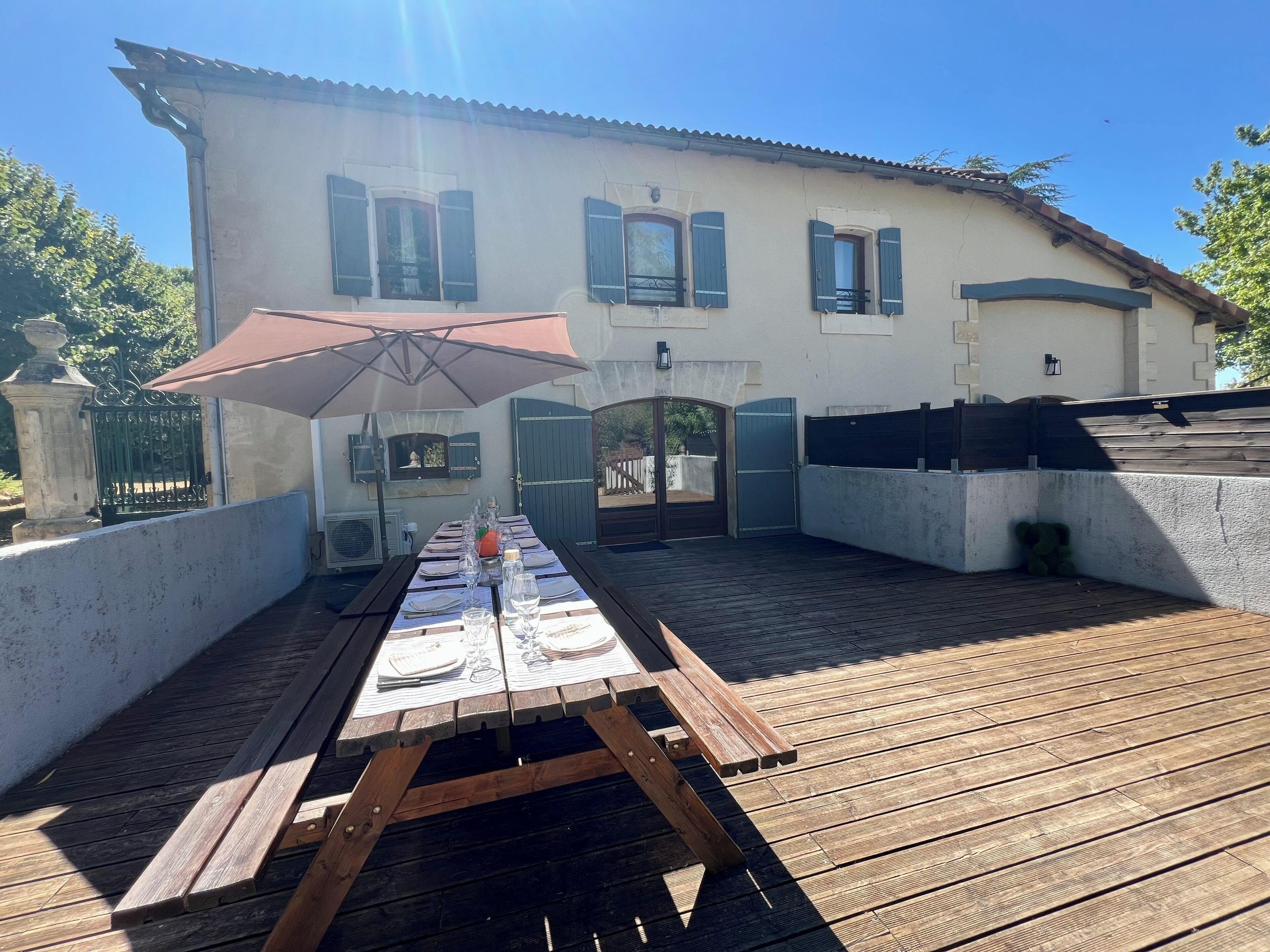 Une terrasse en bois avec une table de pique-nique installée sous un parasol, un bâtiment aux volets bleus et une entrée fermée par un portail.