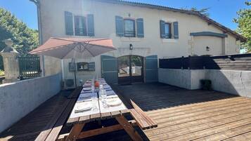 Une terrasse en bois avec une table de pique-nique installée sous un parasol, un bâtiment aux volets bleus et une entrée fermée par un portail.