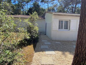 A small, single-story building with a tiled roof, a narrow pathway leading to a door, and a tree in the foreground.