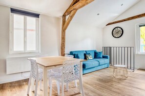 Um quarto com uma mesa de jantar de madeira, cadeiras brancas, um sofá azul, um relógio redondo e uma janela com persianas.