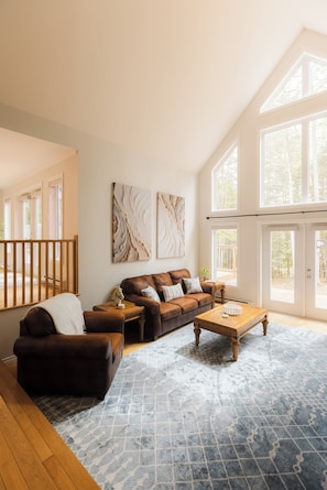 A spacious living room with a large window, a brown leather sofa, a wooden coffee table, and a patterned rug.