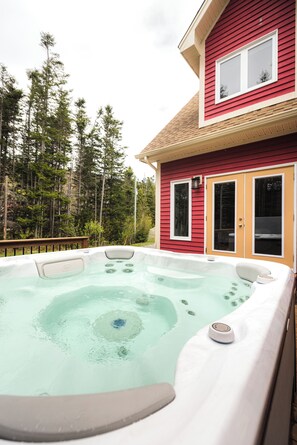 Un bain à remous devant une maison rouge, avec une terrasse en bois et une forêt en arrière-plan.