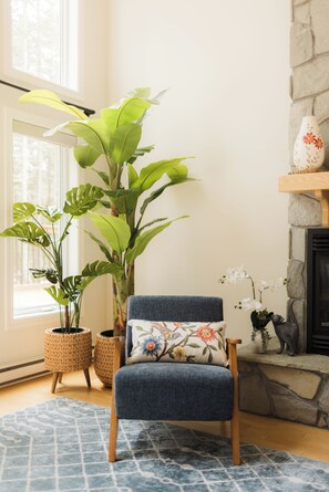 Une pièce avec une grande plante en pot, une chaise bleue agrémentée de coussins à motifs floraux, une cheminée en pierre et un panier en osier.