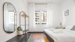 Habitación de hotel con cama, espejo, planta, perchero y vistas a edificios a través de la ventana.