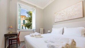 A hotel room with a bed, a desk, a chair, a window with a view, and a painting on the wall. - Beach | 2BR| Luxury Genoa| Wifi By Tilo (Genoa)